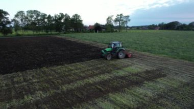 Bir traktör kırsal bir alanda tarlayı sürerek toprağı ekime hazırlıyor. Güneş arka planda batıyor..