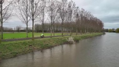 Tek bir bisikletçi bulutlu bir günde, yaprakları olmayan ağaçlarla çevrili bir kanalda yol alır..