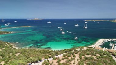 Sardunya kıyı şeridinin çarpıcı manzarası, kristal berrak turkuaz suları, beyaz bir kumsal, yemyeşil bitki örtüsü, ve kayıklarla dolu bir liman..