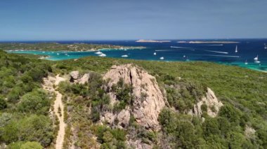 Sardunya 'da turkuaz bir körfeze bakan kayalık manzaralı, parlayan sularda benekli sayısız yelkenli..