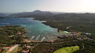 Sardinya 'da sakin bir marinanın panoramik görüntüsü, kıyı şeridi boyunca uzanan kayıklar, yemyeşil ve parlak mavi sularla çevrili..