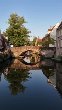 Bruges, Belçika 'da sakin bir kanalın üzerinde bir köprü var. Su, binaları ve yemyeşil yaprakları yansıtıyor. Güzel ve huzurlu bir manzara yaratıyor..