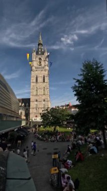 Gent Kuleleri 'nin çan kulesi turist ve yöre sakinlerinden oluşan hareketli bir kalabalığın üzerinde, parlak güneş tarafından mavi gökyüzü zeminine karşı aydınlatılıyor.