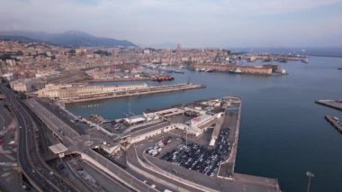 A breathtaking view of Genova showcases the bustling harbor, with ships docked and the cityscape stretching across the shoreline under a clear sky.