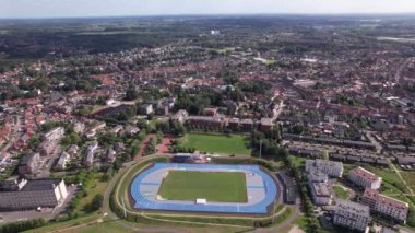 Canlı bir kasabada yuva yapmış, açık gökyüzünün altında parlak mavi izler ve yemyeşil alanlar gösteren geniş bir spor stadyumu keşfedin..