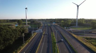 Otomobiller, batan güneşin sıcacık parıltısıyla aydınlatılan yeşillik ve rüzgar türbinleriyle dolu bir otoyol boyunca seyahat ederler..