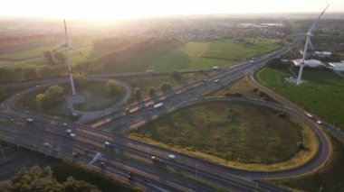 Arabalar, güneş batarken otoyolda sabit bir şekilde dolaşarak manzaranın ve yakınlardaki rüzgar türbinlerinin resimli bir görüntüsünü oluşturuyor..