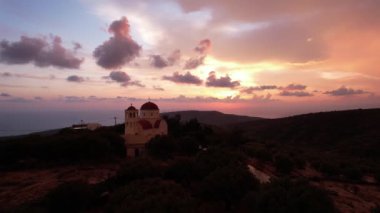 Güneş Girit 'in pitoresk tepelerinin ardında batarken, huzurlu manzarayı ve manastırı aydınlatan gökyüzünün büyülü dönüşümüne tanık olun..