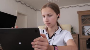 Genç bir kız evde oturmuş, tabletine odaklanmış, kulaklık takmış ve favori oyunlarının tadını çıkarıyor..