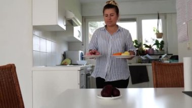 Bir kadın, iyi aydınlatılmış bir mutfakta sağlıklı bir yemek hazırlarken bir tabak taze meyve ve sebze taşır..