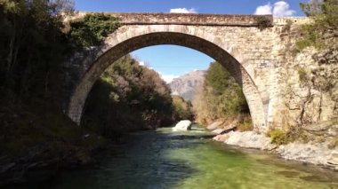 Bereketli yeşillik ve dağlarla çevrili sakin bir nehri kaplayan tarihi bir taş köprünün çarpıcı manzarası..