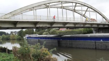 Büyük bir mavna bulutlu bir günde yemyeşil yemyeşil ile çevrili sakin bir nehrin üzerindeki köprünün altında yavaşça hareket eder..