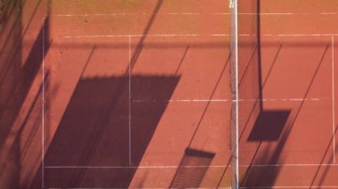 Öğleden sonra yapılan bir tenis antrenmanı, ağ gölgeleri ve kil kortunun çevresini göstererek sakin bir atmosfer yaratıyor..