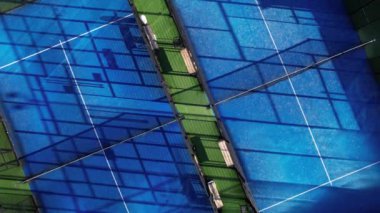 Tenis kortları canlı mavi yüzeyler ve gün ortası güneşinin altında etkin bir spor ortamını ön plana çıkaran güçlü gölgelerle sergilenir..