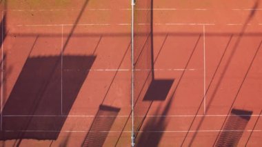 Sıcak bir gün batımının altında canlı bir kil kortunda tenis ağları tarafından oluşturulan gölgelerin etkileşimini deneyimleyin..