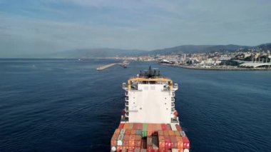 Bir kargo gemisi, canlı konteyner yığınları ve güzel manzaralarla dolu limana doğru sorunsuzca yol alır..