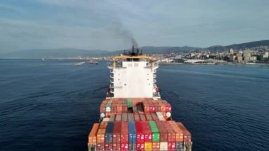 Bir kargo gemisi sakin sularda sorunsuzca ilerler. Arka planda çarpıcı bir şehir silueti vardır..