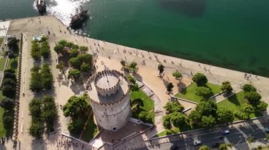 Ziyaretçiler, ikonik Beyaz Kule ve parlak mavi gökyüzünün altındaki yemyeşil park alanlarına bakarak Selanik kıyısı boyunca sakin bir gezinti yapıyorlar..