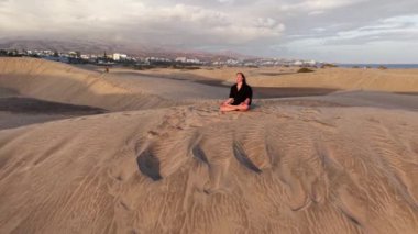 Bir kız, Gran Canaria 'nın kumlu kumullarında huzur içinde oturur ve güneş batarken meditasyon yapar. Alacakaranlığın ılık renkleri kendi yansıması için mükemmel sakin bir atmosfer yaratır..