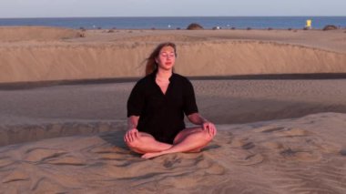 Gran Canaria 'nın yumuşak kumlarında bağdaş kurmuş bir kız batan güneşin sakinleştirici enerjisini emiyor. Altın ışık, okyanus kenarındaki huzurlu meditasyon deneyimini güçlendirir..
