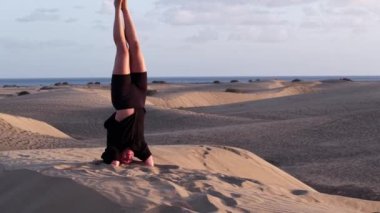 Gran Canaria 'nın sakin kumullarında, bir kız yoga yeteneklerini amuda kalkarak gösteriyor. Huzurlu manzara ve yumuşak okyanus esintisi dinlenmek ve odaklanmak için sakin bir atmosfer yaratır..