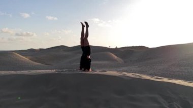 Bir kız, güneş batarken Gran Canaria 'nın büyüleyici kumullarında yoga pozu veriyor. Huzurlu atmosfer ve güzel manzara rahatlama ve farkındalık için ideal bir ortam yaratır..