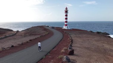 Bir adam, Gran Canaria 'da manzaralı bir deniz fenerine giden sahil yolunda huzurlu bir yürüyüşün tadını çıkarıyor. Güzel manzara okyanusu ve kayalık manzarayı gözler önüne seriyor..