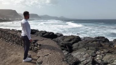 Sakin bir öğleden sonra, bir adam Gran Canaria 'nın kayalık sahilinde okyanusun çarpan dalgalarını gözlemliyor. Güzel manzara düşünmek için huzurlu bir atmosfer yaratır..