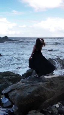 Dalgalı siyah bir elbise giyen bir kız Tenerife 'nin kayalık kıyı şeridinde duruyor ve berrak bir gökyüzünün altındaki okyanus dalgalarını seyrediyor..