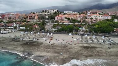 Tenerife 'nin çarpıcı bir hava perspektifini keşfedin. Şemsiyelerle benekli güzel bir plajı ve tatil köyleri ve dağların arka planında güneşlenenleri görün..