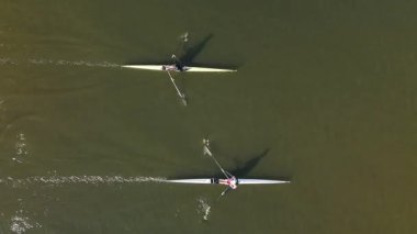 İki sporcu, sakin bir nehirde farklı teknelerde kürek çekerek yeteneklerini sergiliyor ve nazik dalgaların arasına odaklanıyor. Sabahın erken saatleri huzurlu atmosferi güçlendirir..