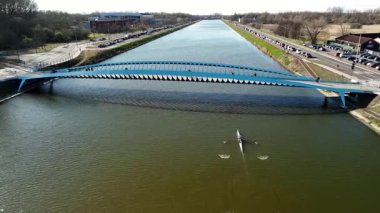 Kürek çeken bir ekip, mimari açıdan kendine özgü mavi bir köprünün altında sakin sularda süzülüyor. Çevredeki manzarada ağaçlar ve arabalar manzaralı yol boyunca uzanır..