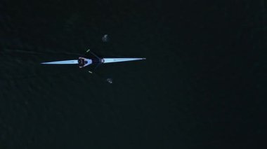 Yalnız bir sporcu sabahın erken saatlerinde parlak, açık mavi bir tekneyle durgun suda kürek çeker. Huzurlu ortam bu spora olan bağlılığı ve odaklanmayı yansıtır..