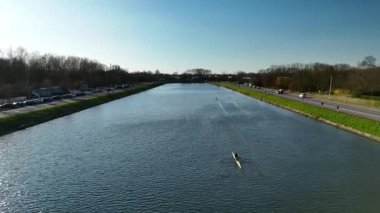 Gün batımında sakin bir nehir sahnesinde sakin sularda süzülen kürek meraklıları görülüyor. Ağaçları çevreleyen ve uzak yollar açık hava sporunun sakin atmosferini arttırır..