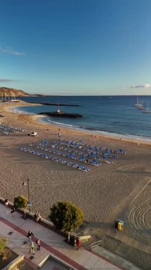 Tenerife 'deki Los Cristianos plajında sakin bir anın tadını çıkarın. Yumuşak kumların renkli bir günbatımı gökyüzü altında hafif dalgalarla buluştuğu yerde. İnsanlar gezinti güvertesinde geziniyor, sakin atmosferi geliştiriyor..
