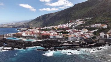 Tenerifes kıyı şeridinin nefes kesici manzarası engebeli dağların ve berrak mavi suların arka planına kurulmuş büyüleyici binaları gösteriyor. Benzersiz manzarayı keşfedin.