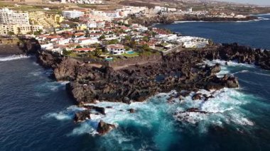 Çarpışan dalgaların engebeli kayalarla buluştuğu Tenerifes kıyılarının nefes kesici manzaralarını keşfedin. Atlantik Okyanusu 'nun arka planında yuva yapmış deniz kenarındaki evlerin güzelliğini görün..