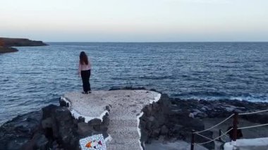 Siyahlı bir kız okyanusa bakan kayalık bir platformda duruyor. Tenerife 'deki sakin dalgaların ve gün batımının tadını çıkarıyor. Huzurlu atmosfer yansıtıcı bir hava yaratır..