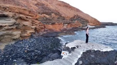 Siyah elbiseli bir kız Tenerife 'nin kayalık kıyılarında duruyor, güneş kayalıkların ardında batarken okyanusa bakıyor, büyüleyici doğal güzellikler ve canlı renklerle çevrili..