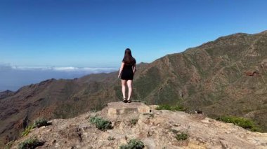 Siyah bir elbiseyle kayalık bir çıkıntıda duran bir kız berrak mavi gökyüzünün altındaki görkemli Tenerife dağlarının nefes kesici panoramik manzarasını seyrediyor..