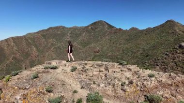 Bir kız siyah elbiseli kayalık bir çıkıntıda duruyor, nefes kesici Tenerife dağlarına bakıyor. Doğanın canlı renkleri ve berrak bir gökyüzü çevrenin güzelliğini arttırır..