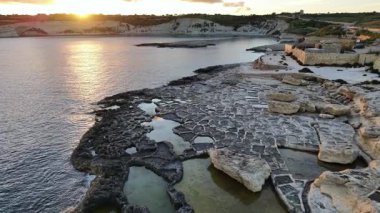 Tanık, Malta kıyı şeridini gün batımı olarak görüyor. Kayalık arazi, sakin sulardaki altın renkleri yansıtır. Doğanın güzelliğini ve sükunetini ortaya çıkarır..