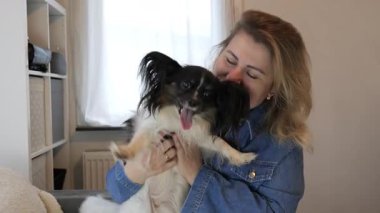Bir kadın küçük köpeğini kapalı bir alanda tutarken gülümsüyor. Ilık ışık, neşeli bir atmosfer yaratıyor ve birlikte eğlenceli bir etkileşim kuruyorlar..