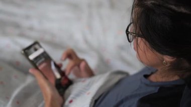 Yatakta rahat bir şekilde yatmış, akıllı bir telefonla meşgul, rahat bir yatak odası atmosferinde battaniyelere sarılı halde akış içeriğini keşfediyor..