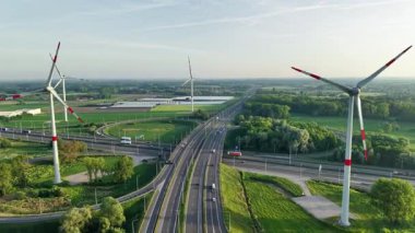 Rüzgâr türbinleri, işlek otoyollara ve geniş alanlara yakın manzaralı kırsal alanlarda dönüyor. Araçlar gün ışığında açık mavi gökyüzünün altında yol boyunca ilerler..