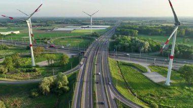 Bruges yakınlarında yemyeşil ve uzun yel değirmenleriyle çevrili dinamik bir yol kavşağına dikkat edin. Altyapı, yenilenebilir enerji ve ulaşım akışının vurgulandığı manzarayla birleşir.
