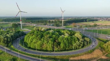 Yeşillik, virajlı yollar ve hareket halindeki araçlarla çevrili dairesel bir alan oluşturuyor. Rüzgâr türbinleri arka planda dik durur ve sakin kırsal atmosferi güçlendirir..