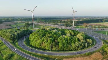 Bu hava perspektifi, açık bir gökyüzüne karşı tarlalar ve rüzgar türbinleriyle çevrili yeşillik ile dolambaçlı bir bereket yakalar. Dairesel yolun etrafında trafik örgü örüyor..