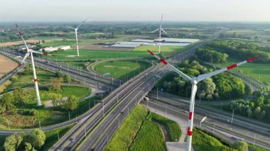 Rüzgâr türbinleri, kırsal alanlarda temiz enerji üretimini gösteren otoyol ve tarlaların yanında dimdik duruyor. Verimli yeşil manzara doğa ve teknoloji arasındaki uyumu vurguluyor..