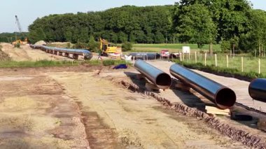 İnşaat işçileri, ağaçlarla çevrili bir kırsal alanda doğalgaz boru hattının bölümlerini birleştirirken görülüyor. Takım çalışmasını vurgulayarak, ağır makineler kurulum sürecinde aktif olarak yer alır.